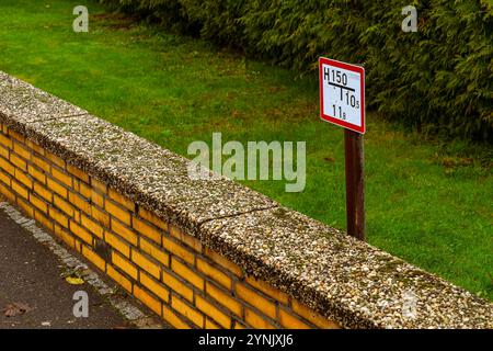 Neben einer strukturierten Wand und einer grasbewachsenen Landschaft befindet sich ein Warnschild mit Höhenbeschränkungen und zeitlichen Beschränkungen in ruhiger Umgebung im Freien. Stockfoto