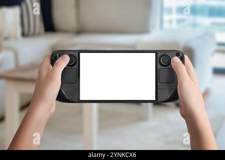 Flache Lay- oder Vorderansicht der Hände, die eine schwarze Handspielkonsole mit isoliertem Bildschirm halten, gemütlicher Hintergrund im Wohnzimmer, ideal für Mockup-Spiele Stockfoto