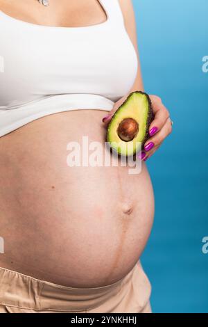 Nahaufnahme einer schwangeren Frau, die eine Avocado vor ihrem Bauch hält, was die Ernährung und die Gesundheit der Schwangerschaft symbolisiert Stockfoto