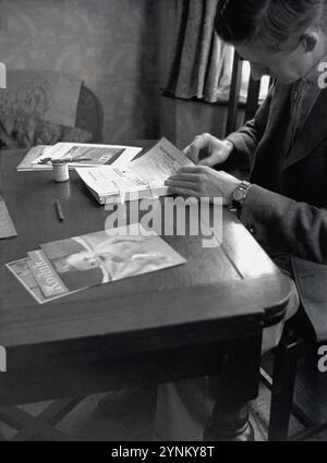 1950er Jahre, Geschichte, Amateurfotografie, in einem Raum, an einem kleinen Holztisch sitzend, ein Mann in einer Sportjacke, Hemd und Krawatte, mit Baumwollfaden, eine Reihe kleiner Fotozeitschriften, England, Großbritannien. Der erste Titel, der 1884 für den Fotografen veröffentlicht wurde, war Amateurfotograf. Im Laufe der folgenden Jahrzehnte wurden andere Publikationen veröffentlicht, unter anderem von Produzenten von Kamerafilmen, wie Kodak, dessen britische Firma eine Zeitschrift, Kodak Magazine, veröffentlichte, ursprünglich das Kodak Magazine for Amateur Photographers. Stockfoto