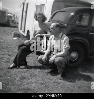 1950er Jahre, historisch, eine junge Frau, in Hosen (Hosen) und mit einem kleinen weichen Ball, gestützt auf den Kofferraum eines Autos der Epoche auf einem Feld vor einer Urlaubskarawane, mit ihrem Vater neben ihr, England, Großbritannien. Auf der Rückseite des Autos befindet sich ein L-Schild für RAC-Fahrlehrer, ein L-Schild aus Metall (Fahrlernschild) mit der Zulassungsnummer des Fahrlehrers. 1935 wurden Fahrkompetenzprüfungen eingeführt, und von dieser Zeit an mussten „L“-Schilder auf Autos angebracht werden, bis die Prüfung abgelegt und bestanden wurde. Stockfoto