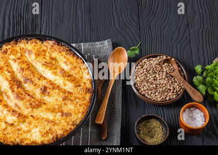 Veganer Hirtenkuchen in Backform auf schwarzem Holztisch mit Zutaten, Gewürzen, frischem Koriander und Holzlöffeln, horizontaler Blick von oben Stockfoto