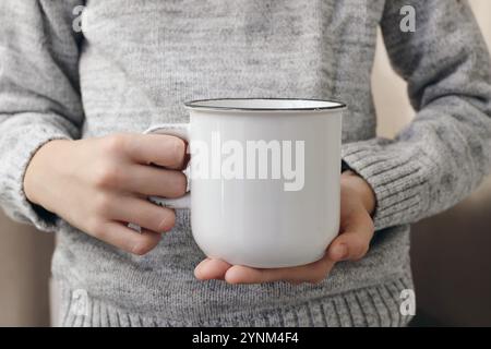 Nahaufnahme der Hände, Finger, die weiße Tasse Tee halten. Leeres Modell des Cearmic Bechers. Unscharfer Hintergrund, selektiver Fokus. Kaffeezeit. Winterstilleben Stockfoto