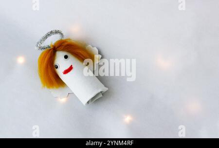 Ein Engel mit langen gelben Haaren, hergestellt von einem Kind aus Toilettenpapierrollen auf weißem Hintergrund mit Weihnachtslichtern. Stockfoto