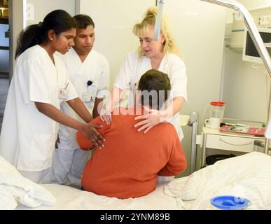 PRODUKTION - 21. November 2024, Sachsen-Anhalt, Halle (Saale): Auf der Station der Universitätsklinik und Poliklinik für Neurologie und Neurochirurgie erklärt die Praxislehrerin Nicole Schmidt (r) den angehenden indischen Krankenschwestern Binny James (l) und Adeesh Krishnan Kutty Sarayu (2. V. l.), wie man einem Patienten nach einer Operation beim Liegen hilft. Sie gehören zu den ersten 16 Auszubildenden aus Indien, die im Ausbildungszentrum für Gesundheitsberufe am Universitätsspital ihre zweite Ausbildung in allgemeiner Krankenpflege und damit in der Betreuung von Menschen jeden Alters in allen Pflegebereichen erhalten. In Stockfoto