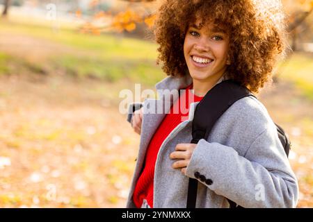 In einem wunderschönen Herbstpark zeigt eine fröhliche junge Frau mit lockigen Haaren ihr helles Lächeln, während sie einen stilvollen grauen Mantel und einen roten Pullover trägt Stockfoto