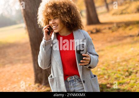 Helles Herbstlaub umgibt eine junge Frau, die lächelt und sich mit einem warmen Getränk in der Hand unterhält. Sie verkörpert einen freudigen Moment Stockfoto