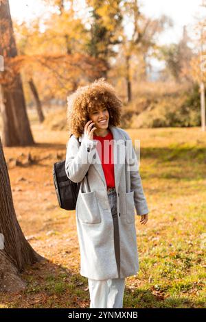 Eine junge Frau mit lockigen Haaren lächelt hell, während sie am Telefon spricht, warm gekleidet in einem stilvollen Mantel inmitten einer malerischen Herbstlandschaft Stockfoto