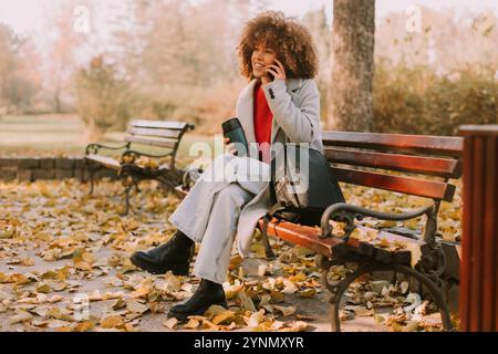 Gemütlich auf einer Bank sitzend, spricht eine fröhliche junge Frau mit lockigen Haaren auf ihrem Telefon, während sie eine Tasse Kaffee genießt, während ich im herbstlichen Herbstlaub i Stockfoto