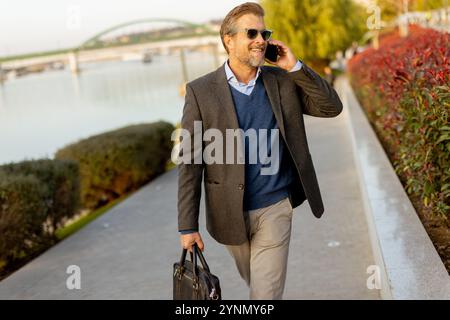 Ein selbstbewusster Geschäftsmann in eleganter Kleidung spaziert neben leuchtenden roten Pflanzen und unterhält sich auf seinem Handy, während die Sonne über dem Wasser untergeht Stockfoto