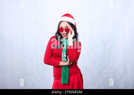 Charmante junge indonesien-Frau mit rotem Pullover mit Weihnachtsmütze und Schal, fröhlich mit der Kamera, die rote Sonnenbrille hält, feiert frohe weihnachten und Stockfoto