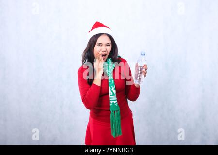 Charmante junge indonesien-Frau mit rotem Weihnachtspullover, Weihnachtsmütze und Schal, schreiend vor die Kamera, während sie Wasser in Flaschen in der linken Hand hält, Isolat Stockfoto