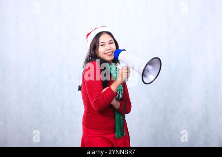 Die junge indonesienfrau trägt einen roten weihnachtspullover, weihnachtsmütze und Schal, ein großes Lächeln auf die Kamerahand, die Megaphon-Lautsprecher und Arm hält, Isolat Stockfoto