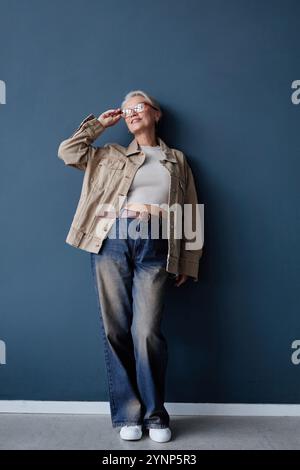 Vertikales Porträt einer modernen Seniorin mit weißem Haar in Brille und lässigem Outfit mit Baggy-Jeans, die im Studio vor der blauen Wand posiert Stockfoto
