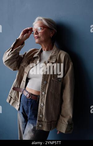 Vertikales Porträt einer modernen älteren Frau mit Brille und lässigem Outfit, die entspannt vor der blauen Wand im Studio mit natürlichem Licht steht Stockfoto