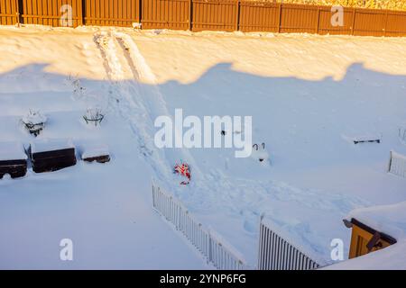 Frisch gefallener Schnee im Hinterhof mit Schlittenbahnen, die bergab führen, umgeben von Holzzaun und Gartenbetten. Schweden. Stockfoto