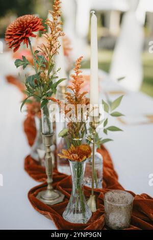 Elegante Einrichtung im Hinterhof mit farbenfrohen Herbstblumen, Messing-Kerzenleuchtern und einer Tischläufer aus Terrakotta-Samt. Stockfoto