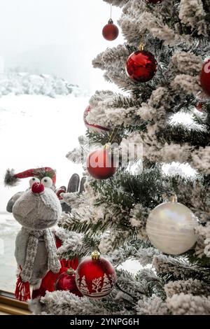 Weihnachtsbaum mit Schneebästen, Ballschmuck und Figur des niedlichen Rentierspielzeugs. Fenster mit Blick auf den Winter Stockfoto