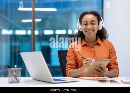 Lächelnde Frau, die Kopfhörer trägt, Notizen auf einem Notizblock am Schreibtisch mit Laptop macht. Ambiance zeigt Positivität, Professionalität, Fokus in Moderne Stockfoto