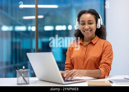 Junge Frau, die Laptop benutzt, Kopfhörer im Büro trägt. Professionell, konzentriert auf die Aufgabe, lächelnd mit Selbstvertrauen. Arbeiten, Produktivität und moderne Arbeitsumgebung demonstrieren. Stockfoto
