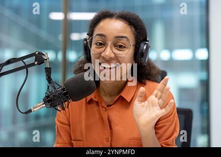 Lächelnde junge Frau mit Mikrofon und Kopfhörern für Podcasts im Studio. Sie winkt selbstbewusst und schafft eine warme, einnehmende Atmosphäre. Ideal für Themen Stockfoto