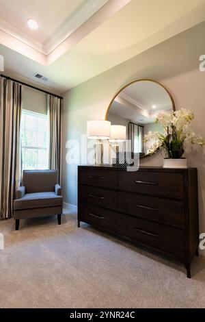 Elegante Schlafzimmerecke mit dunklem Holzkommode, rundem Spiegel, gemütlichem Sessel und natürlichem Licht, das eine warme und moderne Ästhetik ausstrahlt. Stockfoto