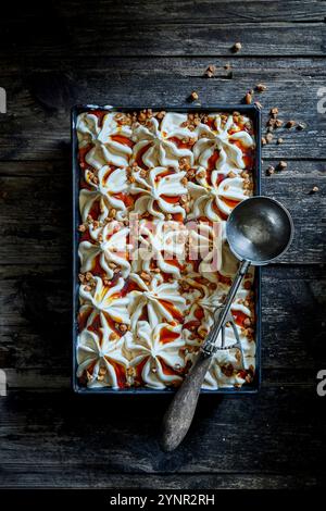 Hausgemachtes Karamelleis mit Karamellsauce, karamelisierten Nussstückchen, in einer Backform auf dunklem hölzernem Hintergrund mit einem alten Eislöffel Stockfoto