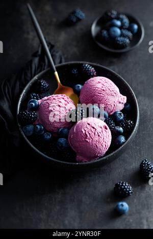 Drei Bällchen brombeereis in einer schwarzen Schüssel mit Heidelbeeren, Brombeeren und einem goldenen Löffel auf einer schwarzen Oberfläche angeordnet Stockfoto