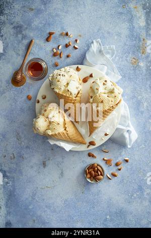 Mandel-Honig-Eis in drei knusprigen Eiskegeln zubereitet, auf einer weißen Platte angeordnet, mit Mandeln und Honig auf blauem Hintergrund dekoriert Stockfoto