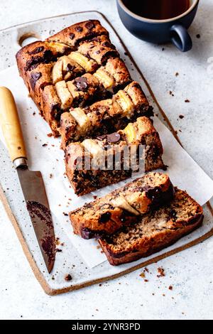 Ein in Scheiben geschnittener Bananenschokoladenbrot auf einem Holzbrett neben einem Messer und einer Tasse Kaffee. Stockfoto