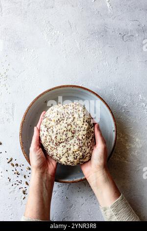 Eine Person, die eine runde Kugel Teig mit Samen in einer Keramikschale auf einer strukturierten Oberfläche hält. Stockfoto