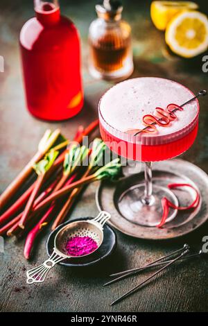 Schöner rosafarbener Cocktail mit schaumiger Oberseite in Coupé-Glas mit einfacher Rhabarber-Curl-Garnitur, frischen Rhabarberstielen und Sirup im Hintergrund Stockfoto