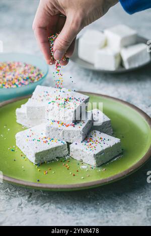 Eine Hand streut bunte Streusel auf einen Stapel quadratischer weißer Marshmallows, die auf einer grünen Keramikplatte gestapelt sind. Eine Schüssel mit weichen Streuseln und Stockfoto