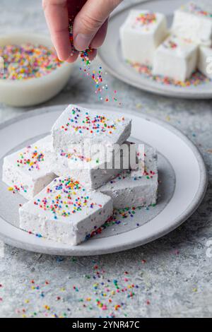 Eine Hand streut bunte Streusel auf einen Stapel quadratischer weißer Marshmallows, die auf einer hellgrauen Platte gestapelt sind. Eine Schüssel mit weichem Fokus und mehr Stockfoto