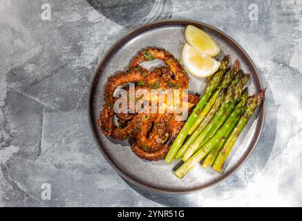 Köstlicher gegrillter Tintenfisch, serviert mit lebhaften Spargelstangen und frischen Zitronenscheiben auf einem grauen Teller. Sonnenlicht-Einmündung, hartes Licht. Stockfoto