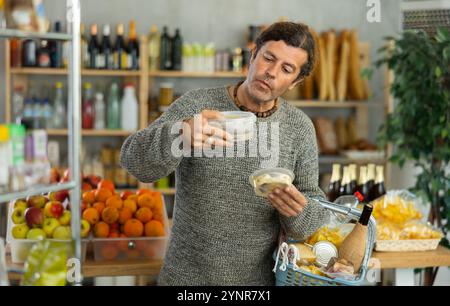 Fokussierter Mann mit Warenkorb, der versucht, eine Wahl zu treffen, der zwei Heringsarten im Supermarkt hält Stockfoto