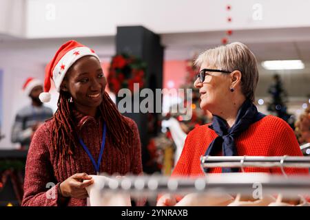 Einzelhandelsmitarbeiter helfen alten Kunden mit Einkaufsratschlägen und bieten Frauen beim Einkaufen von weihnachtsgeschenken Unterstützung. Ladenassistent präsentiert Waren auf Kleiderbügeln für Kunden, festliche Feiertage. Stockfoto