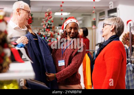 Ein Mitarbeiter im Einzelhandel zeigt alten Kunden einen Blazer und empfiehlt ihnen die perfekten Artikel für weihnachtsessen. Ladenassistentin hilft Kunden beim Kauf formeller Kleidung und präsentiert Waren. Stockfoto