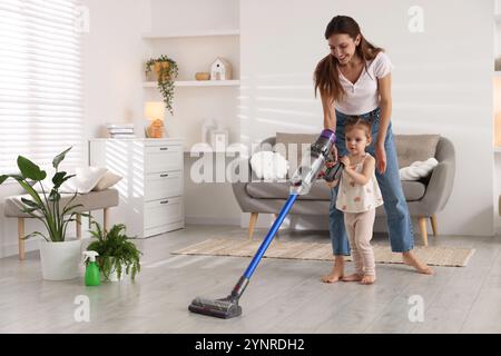 Die lächelnde junge Frau und ihre Tochter putzen zu Hause den Boden mit schnurlosem Staubsauger Stockfoto