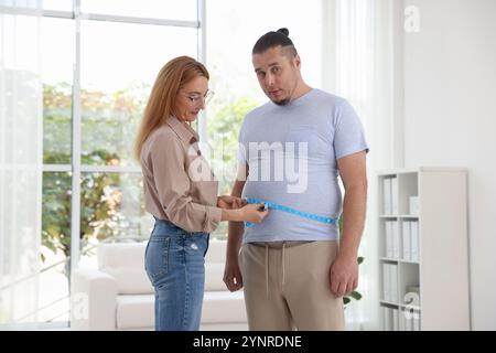 Ernährungswissenschaftler, der die Taille eines übergewichtigen Mannes mit Klebeband in der Klinik misst Stockfoto