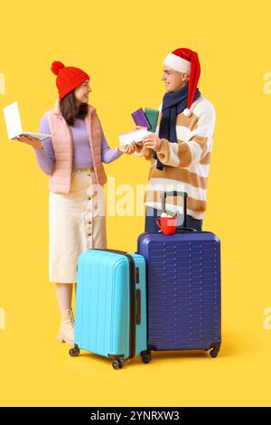 Nettes junges Paar mit Laptop, Pässen und Koffern auf gelbem Hintergrund. Winterurlaubskonzept Stockfoto