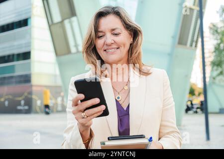 Lächelnde, reife Dame, die im Freien ein Mobiltelefon benutzt, in formeller Kleidung gekleidet, mit einem Notebook in der Hand Multitasking macht und in der Innenstadt immer in Verbindung und produktiv bleibt. Geschäftsfrau, die mit einem Smartphone surft. Hochwertige Fotos Stockfoto