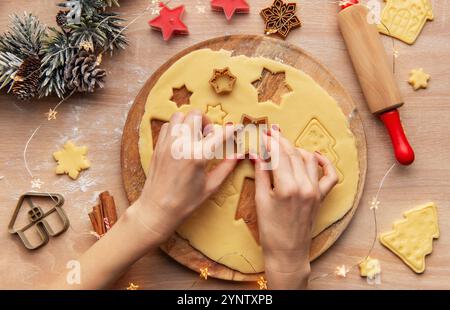 Frau, die weihnachtskekse mit unterschiedlich geformten Keksschneidern auf einem Holzbrett zubereitete, umgeben von festlichen Dekorationen Stockfoto