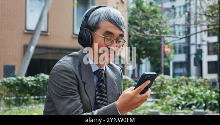 Business, Musik und Telefon mit reifem Mann im Freien in der Stadt für eine Pause vom Job oder der Arbeit. Kommunikation, Kopfhörer und Lächeln mit japanischem Unternehmen Stockfoto