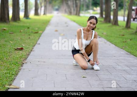 Lächelnde Athletin kniend auf einem Parkweg, bindet ihre Schnürsenkel, bereit zum Training Stockfoto