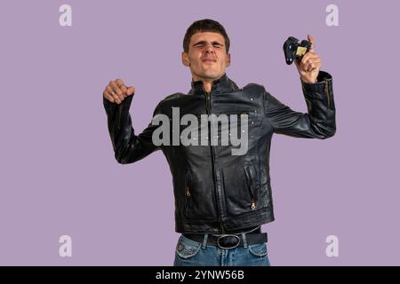 Ein junger Mann in einer schwarzen Lederjacke hebt die Arme und hält ein Gamepad und feiert einen Sieg in einem Videospiel Stockfoto