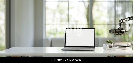 Ein Laptop mit weißem Bildschirm, Kopfhörern und Mikrofon auf einem weißen Tisch in einem modernen hellen Wohnzimmer. Heimradio Podcast Büro. 3D-rendering Stockfoto
