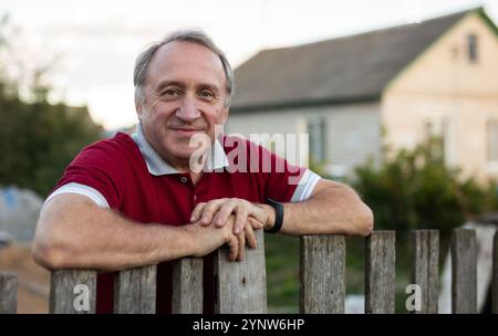 Ein erwachsener Mann, der entspannt neben dem Zaun im Garten steht und lächelt Stockfoto