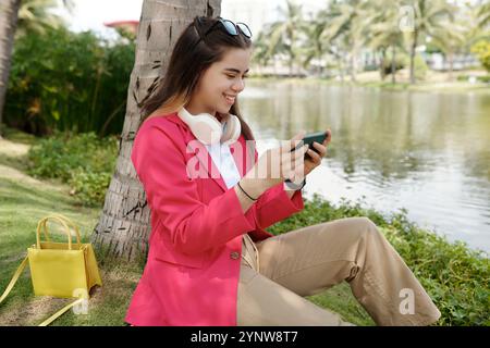 Lächelnde junge Frau, die sich am See entspannt und ihr Smartphone hält Stockfoto