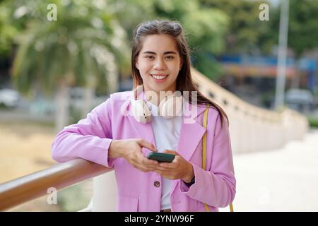 Porträt einer lächelnden jungen Frau, die ihr Smartphone draußen hält Stockfoto